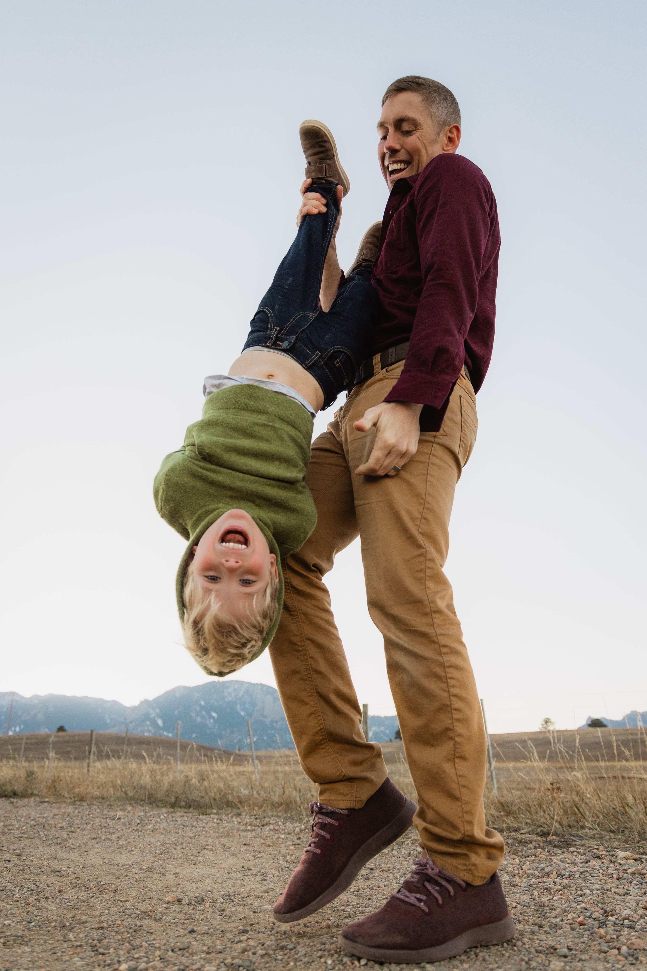Person wearing maroon sweater and khaki pants playfully holds someone upside down in a field with mountains in background.