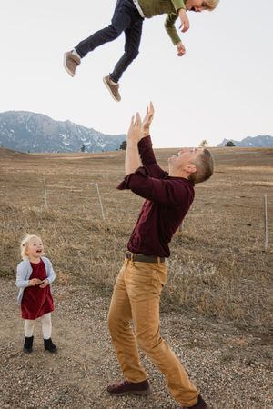 Playful family moment in an open field with mountains, as someone throws a child in the air while another watches.