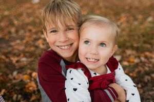 Two children wearing burgundy sweaters smile together while sitting among fallen autumn leaves.