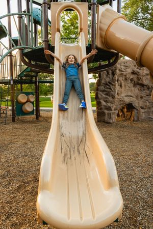 A child enjoying a spiral playground slide on an outdoor playground with climbing structures and a rock wall.