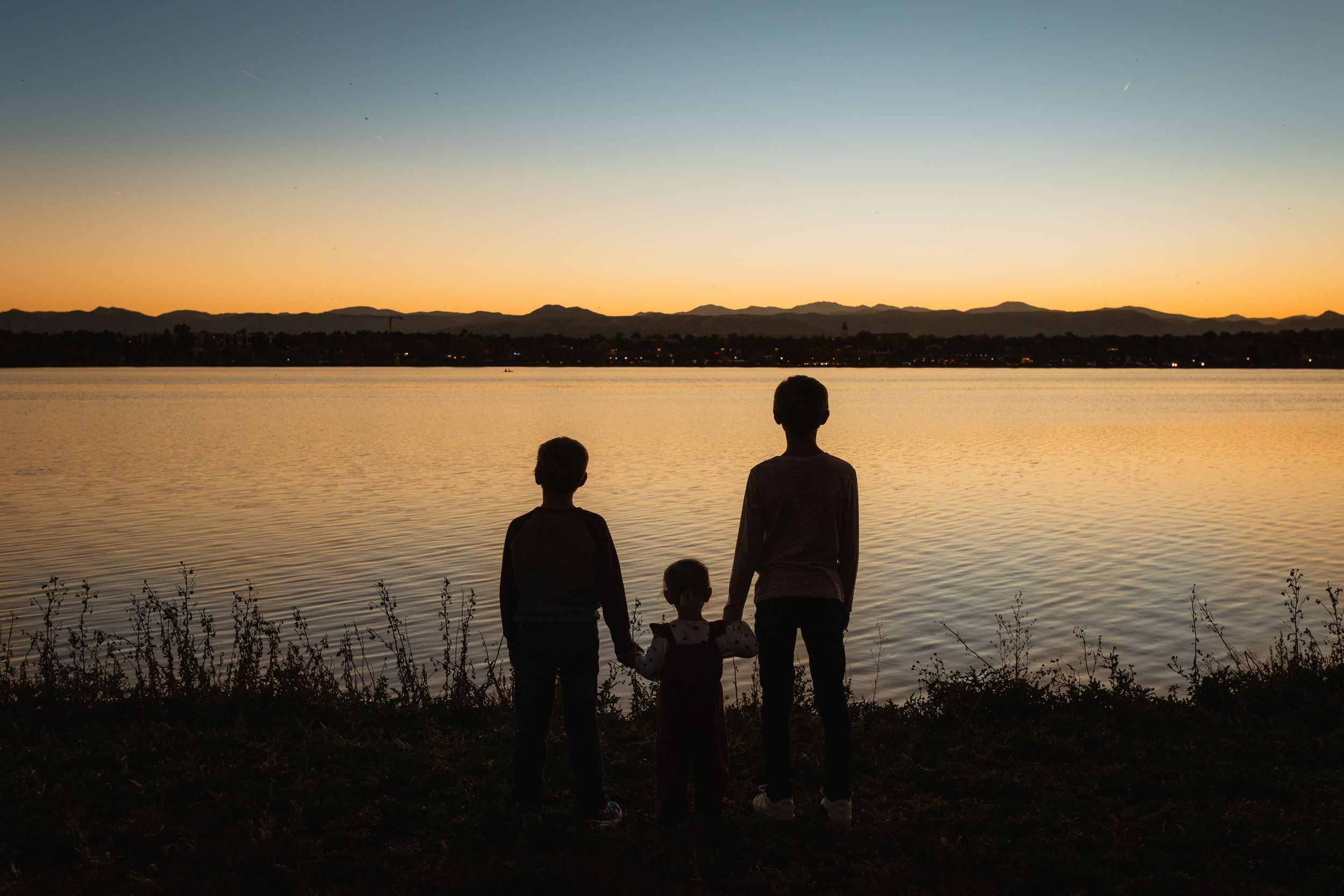 Silhouettes of figures stand on a beach watching a golden sunset over calm water and distant mountains.