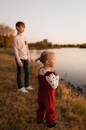 Two children stand on a grassy riverbank at golden hour, with one wearing a red jumpsuit and the other in a light sweater.