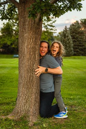 Two people share a joyful, candid moment while sitting against a large tree trunk in a grassy park setting.