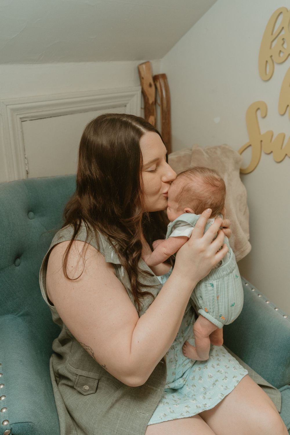 Mother kissing her newborn baby while sitting in a teal velvet nursery chair.