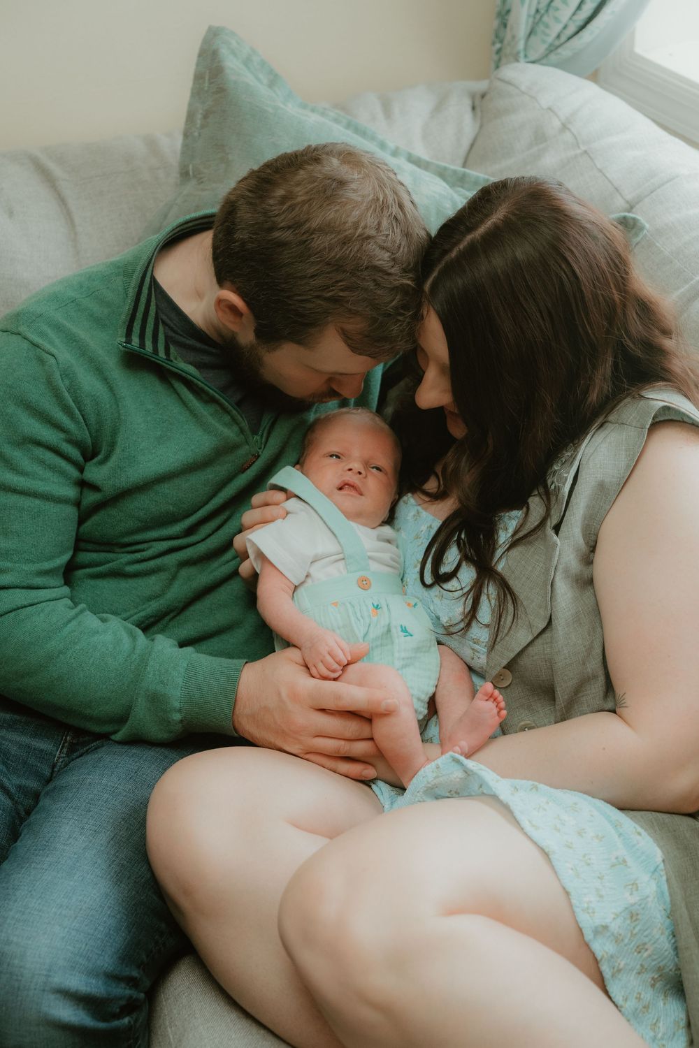 New parents leaning in close together while cradling their smiling newborn baby on a couch.