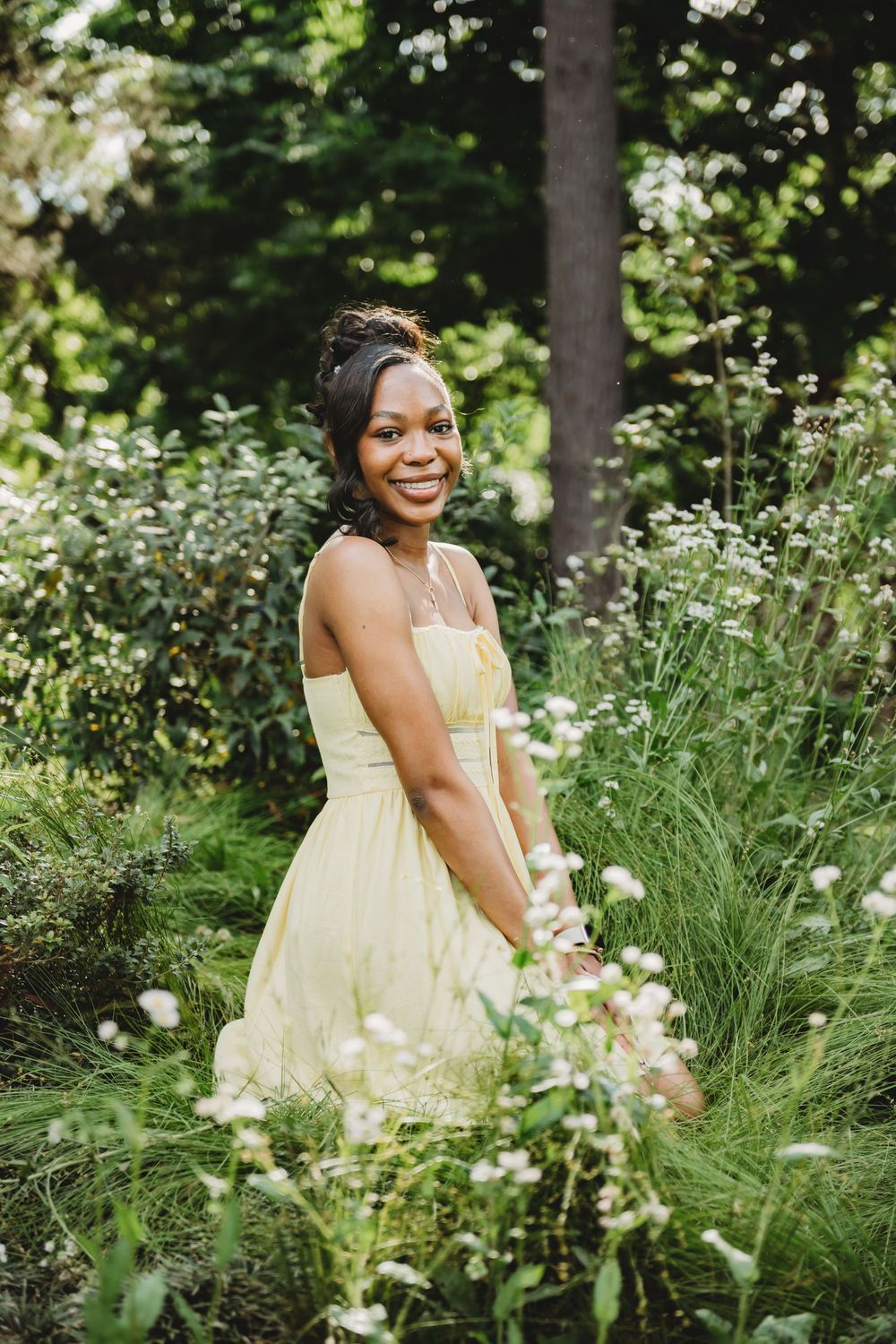 A person in a pale yellow dress poses gracefully among lush garden greenery and white flowers on a summer day.