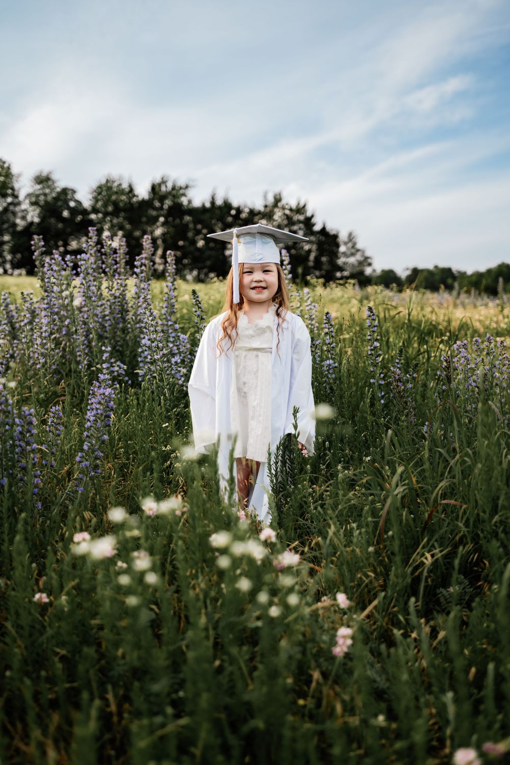 A child in a white graduation cap and gown stands in a wildflower meadow on a sunny day.