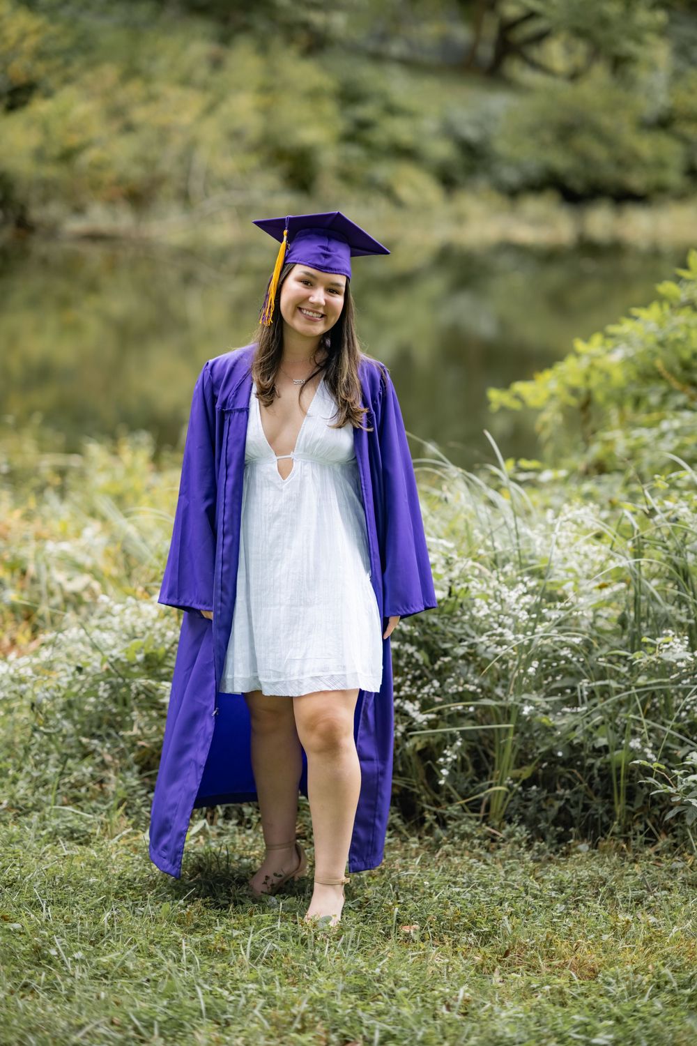 Graduate in purple cap and gown sits in a scenic outdoor setting surrounded by greenery and natural light.