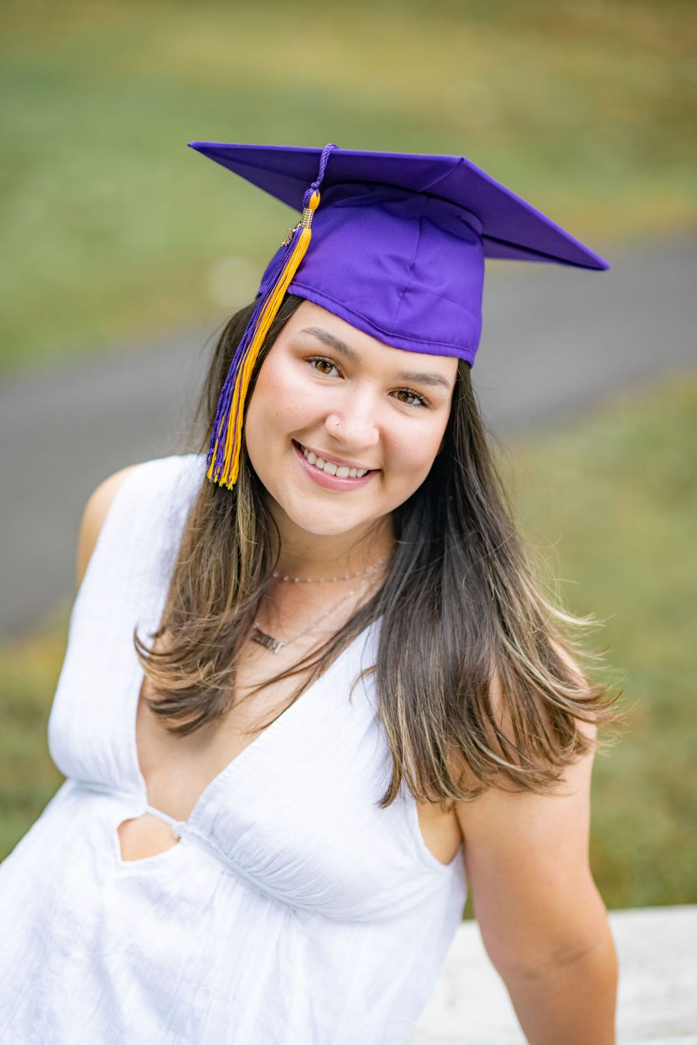 Graduate in white dress wearing purple cap and gold tassel posing outdoors for graduation portrait.