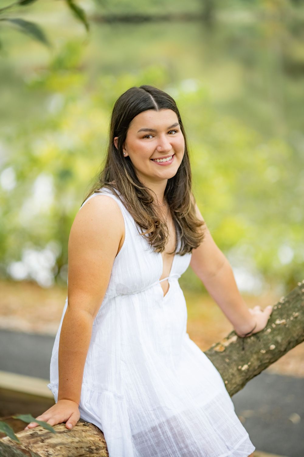 A person in a white summer dress sits on a tree branch outdoors with a warm smile and natural lighting.