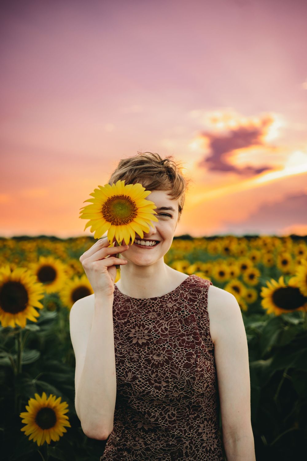 A sunflower field at sunset with a figure in a brown dress playfully holding a sunflower bloom over their eye.