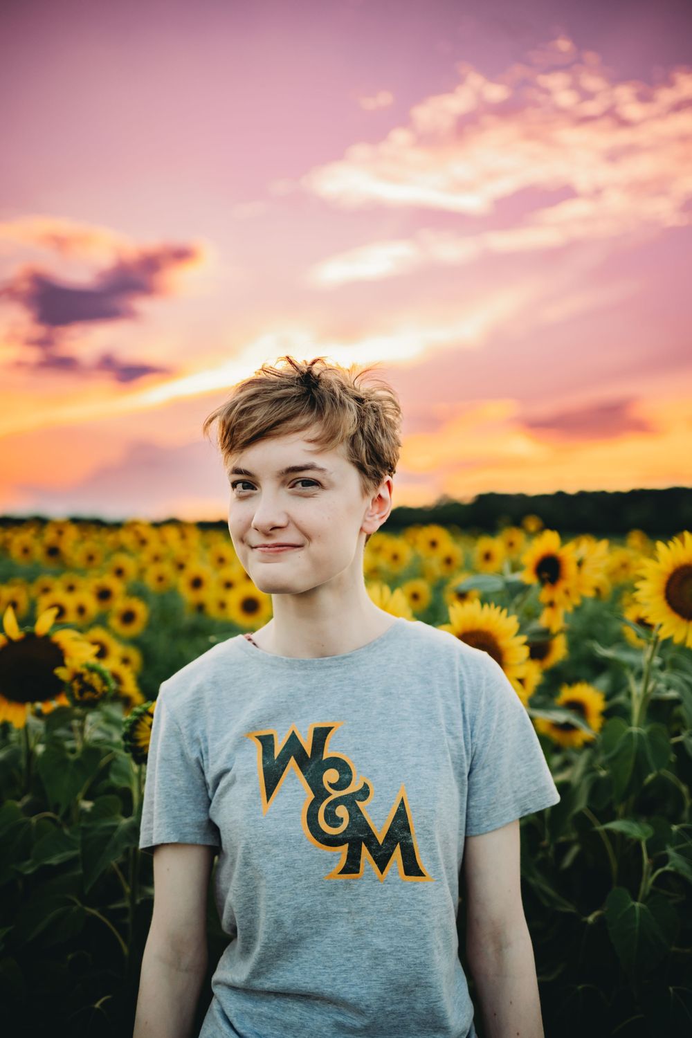 Person wearing a gray t-shirt with W&M text stands in a sunflower field at sunset with pink and orange clouds.