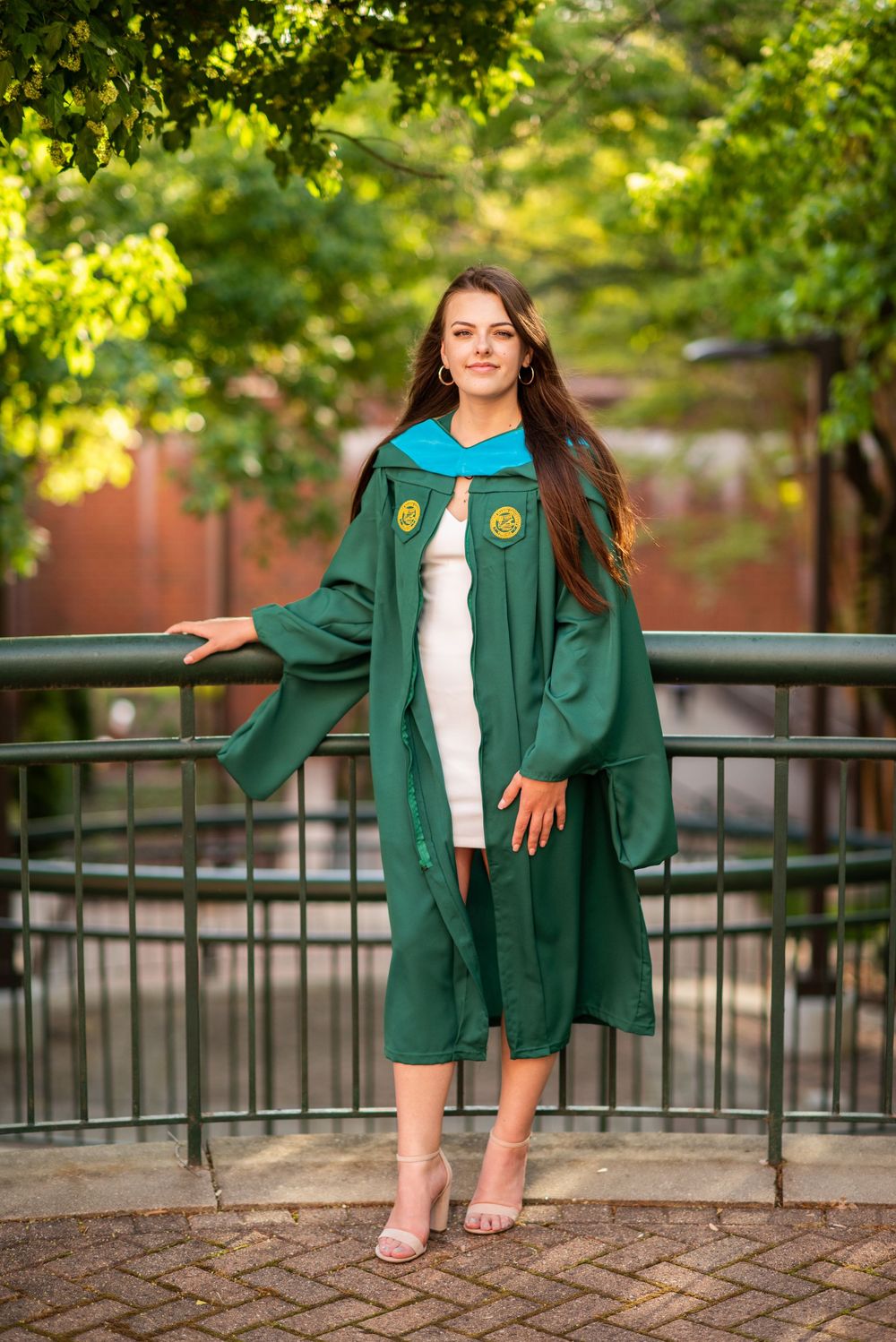 Graduate in green cap and gown poses on brick pathway with iron railing and sunlit foliage in background.