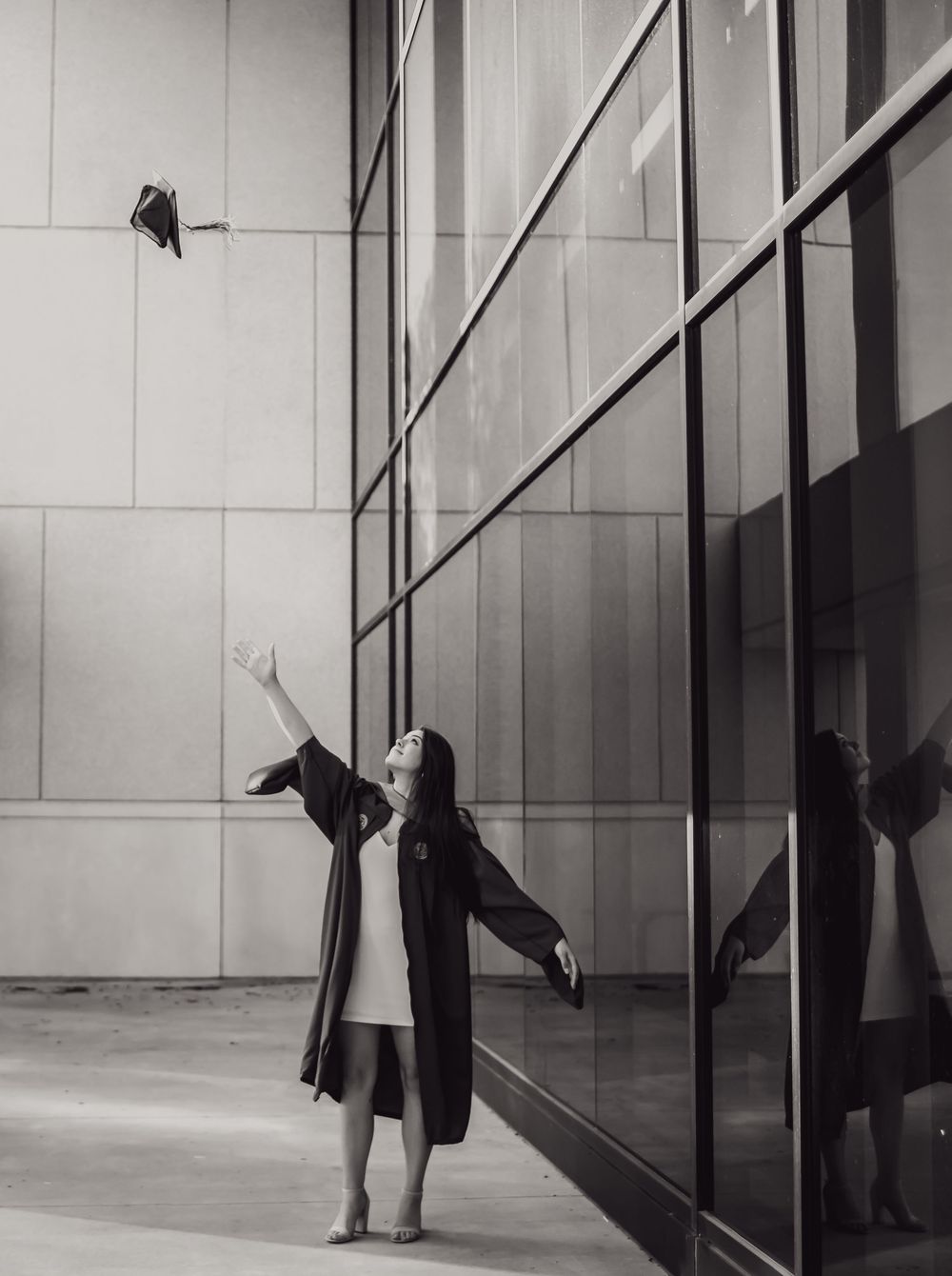 Black and white photo of someone in a long coat reaching up towards the corner of a modern building with reflective glass panels.