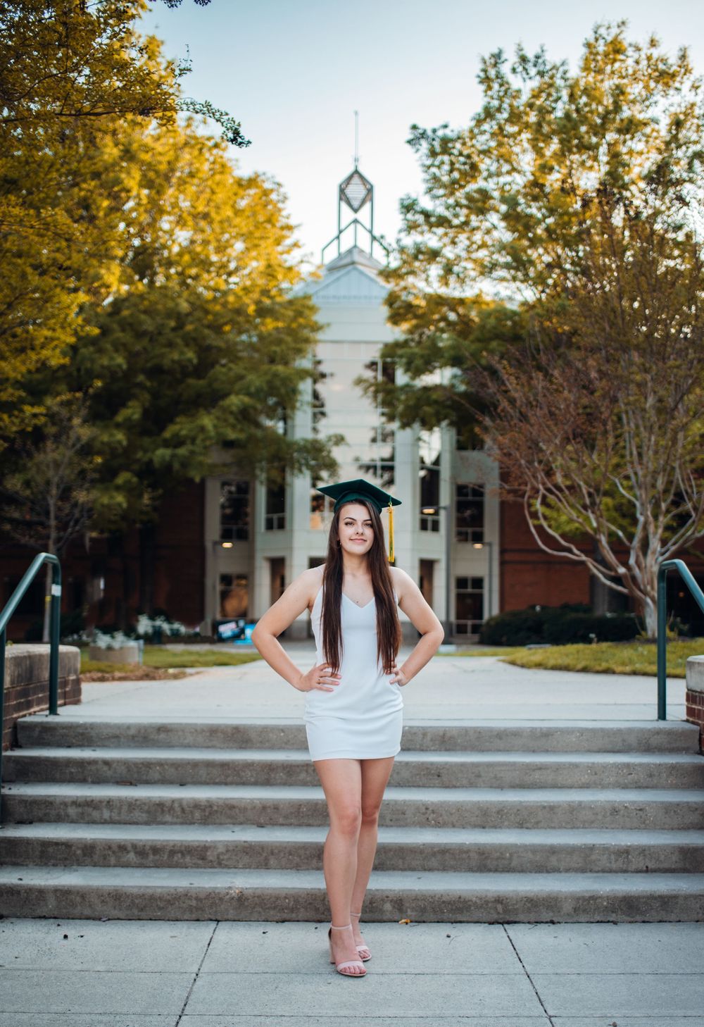 Graduate in white dress poses on steps with a traditional red brick college building and autumn trees in background.