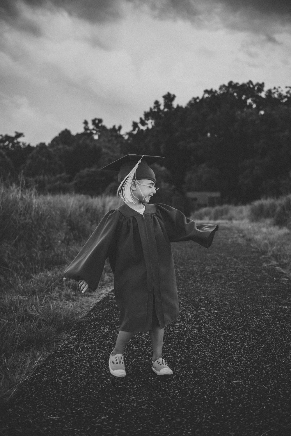 A graduate in black cap and gown stands on a path with arms outstretched against a moody cloudy sky.