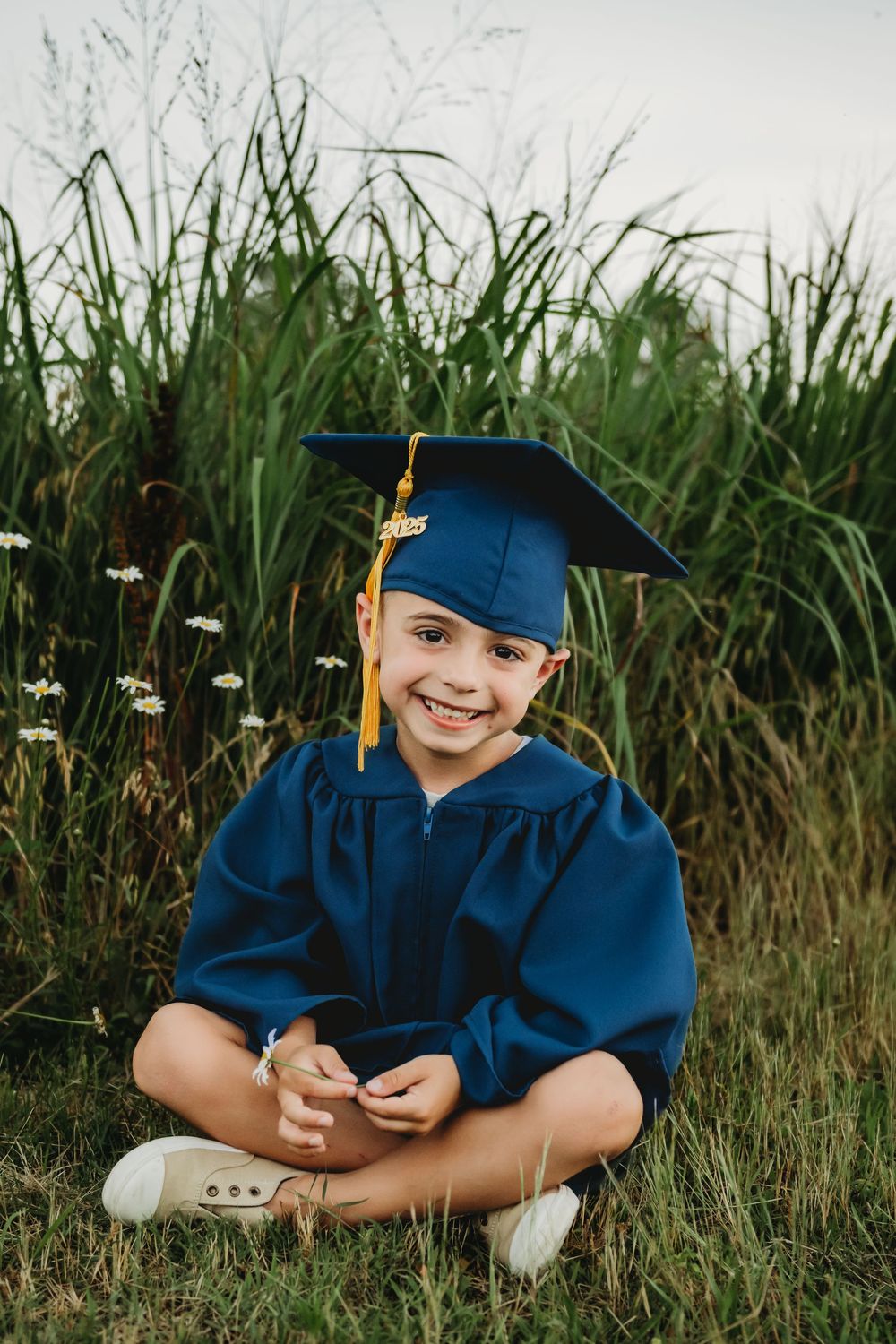 A young graduate in blue cap and gown sits cross-legged in tall grass during a graduation photoshoot.