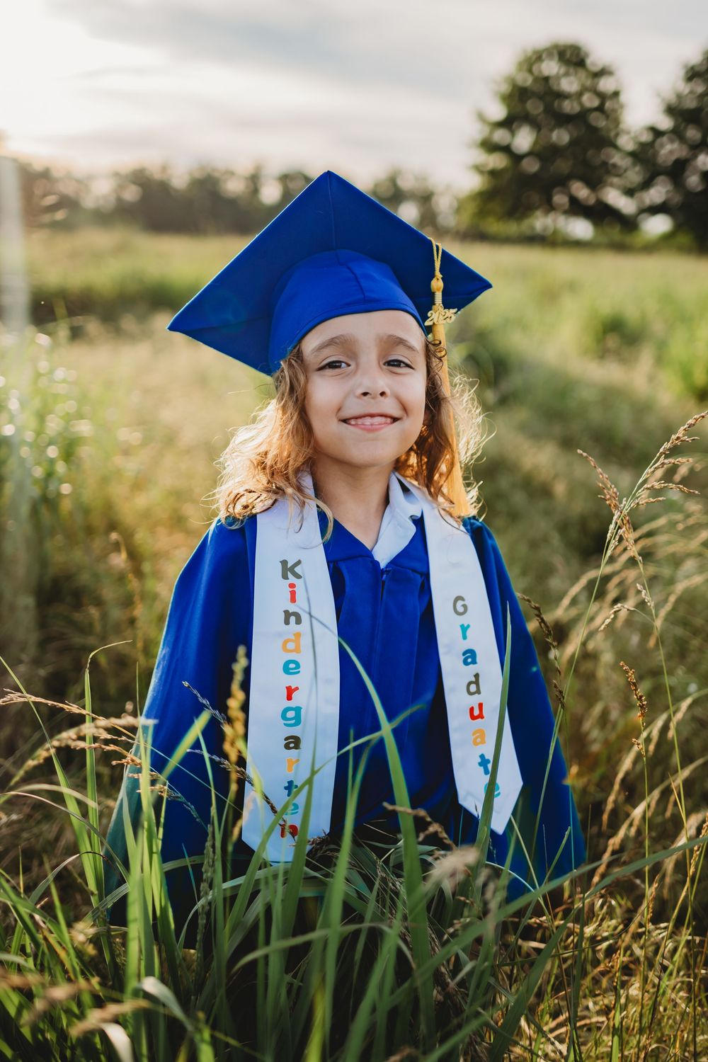 A graduation photo of a young graduate in blue cap and gown with white sash standing in a sunlit field of tall grass.