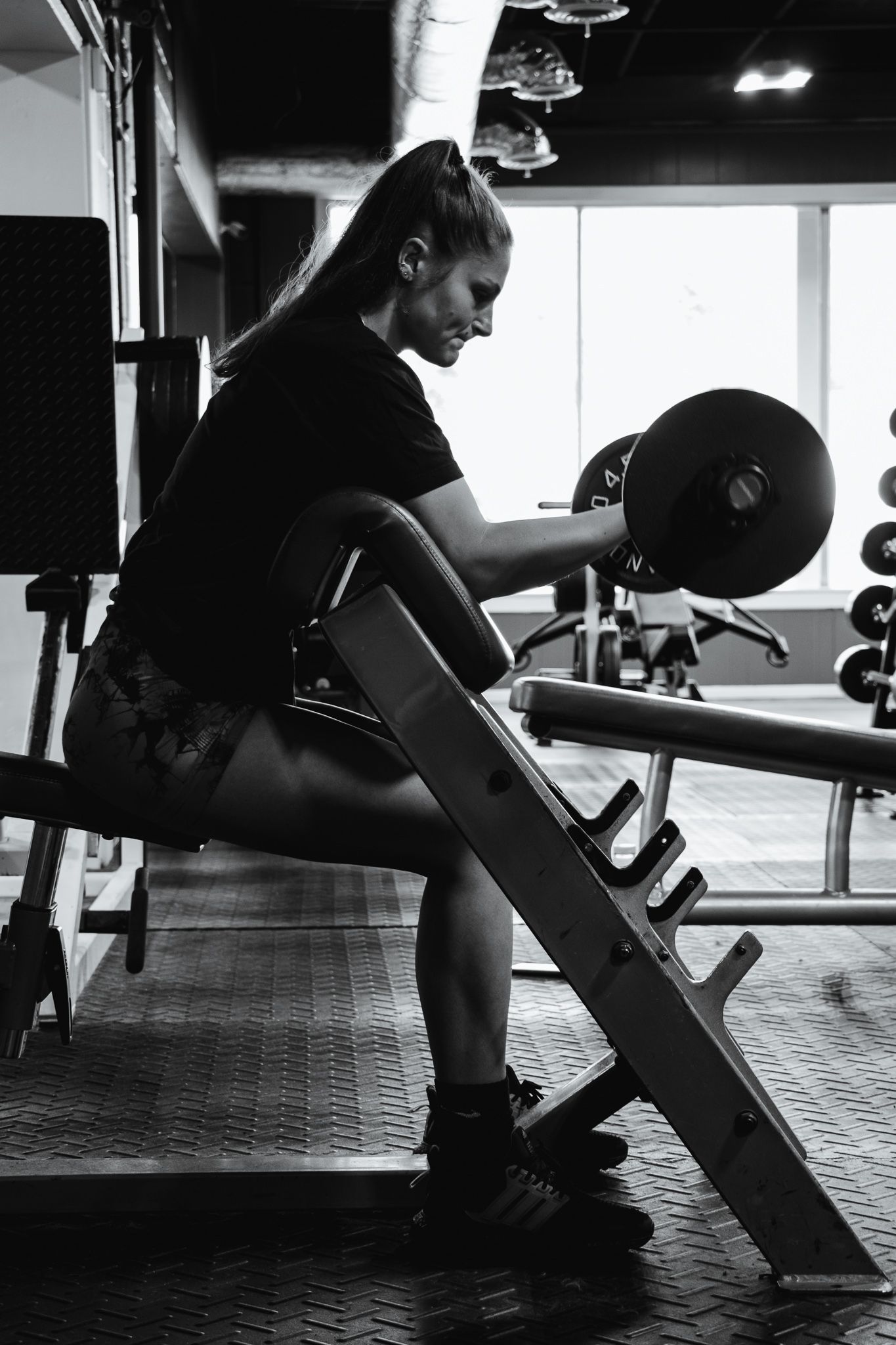 Woman does seated bicep curls with a dumbbell in a gym, silhouetted by light from large front windows.