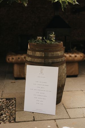 A white paper menu or schedule sits propped against a rustic wooden barrel with decorative plants on top.