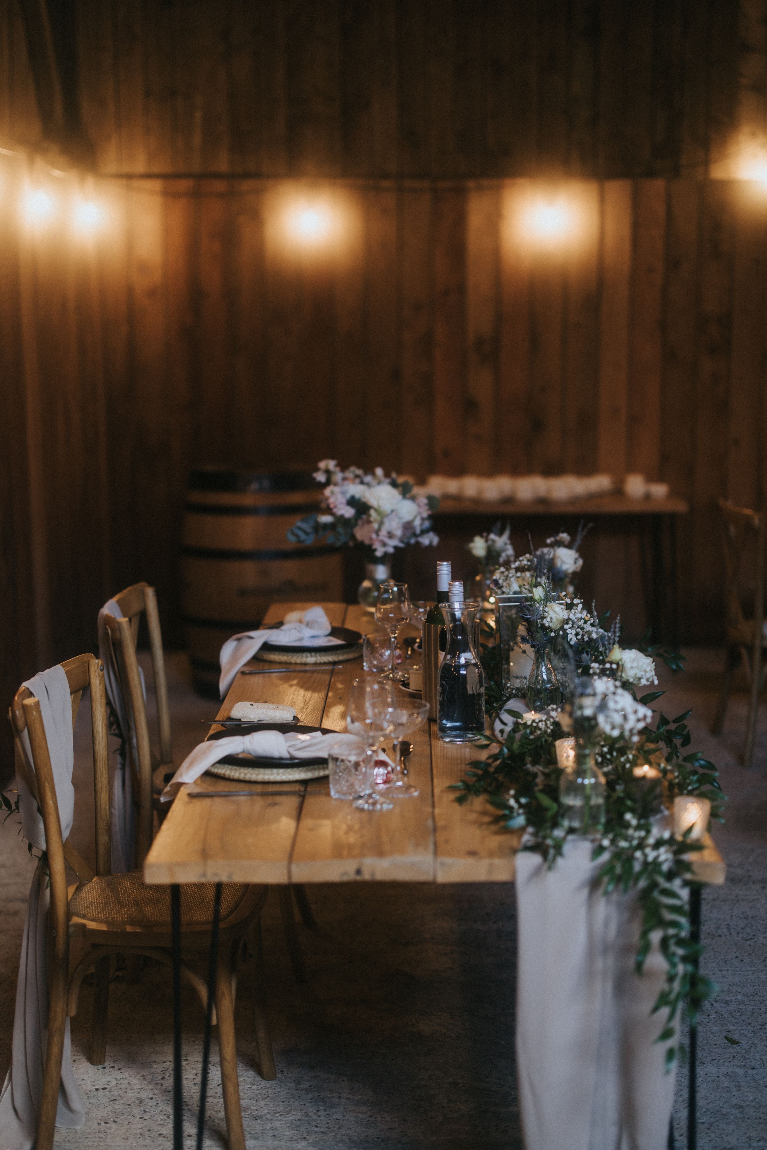 Rustic wooden table set for dinner with string lights and floral decorations in moody barn setting.