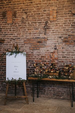Minimalist wedding signage with greenery accents against rustic exposed brick wall and wooden display table.