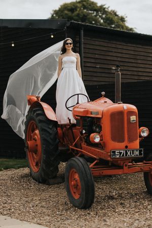 A vintage orange tractor serves as a rustic backdrop for someone in a flowing white wedding dress creating romantic photos.