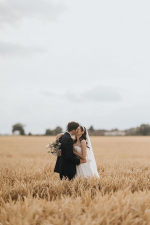 A couple share a romantic moment in a golden wheat field under an overcast sky with bouquet in hand.