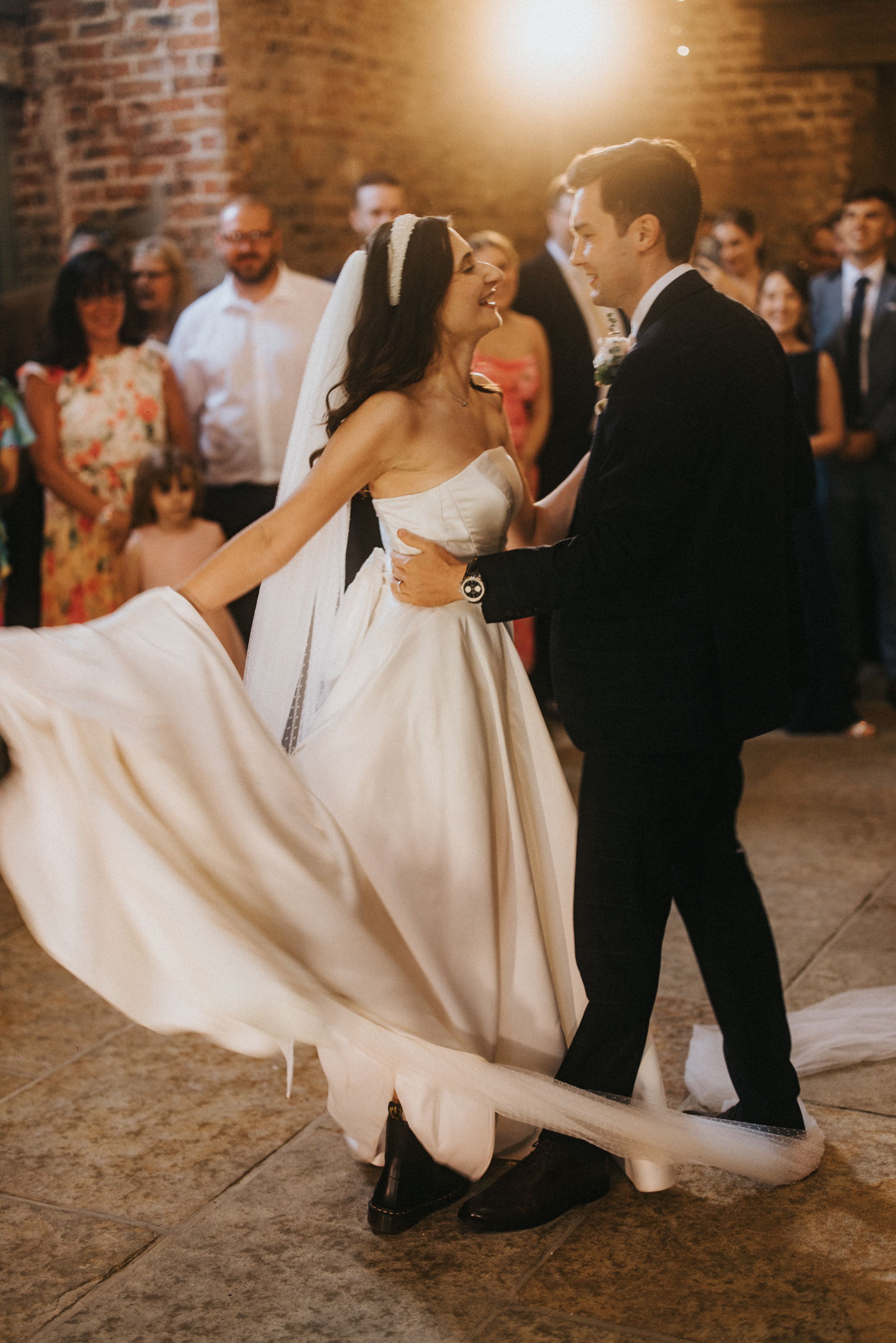 A bride's flowing white wedding dress swirls elegantly during a dance at a rustic brick-walled reception venue.
