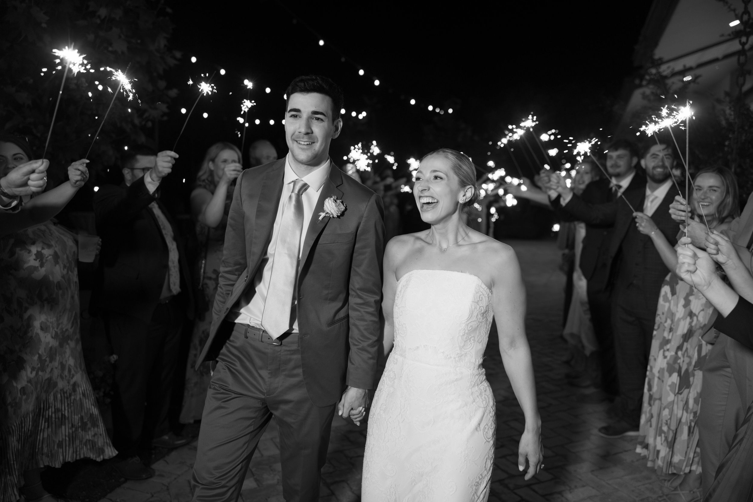 Wedding celebration at night with sparklers glowing as couple walks through guests during outdoor reception.