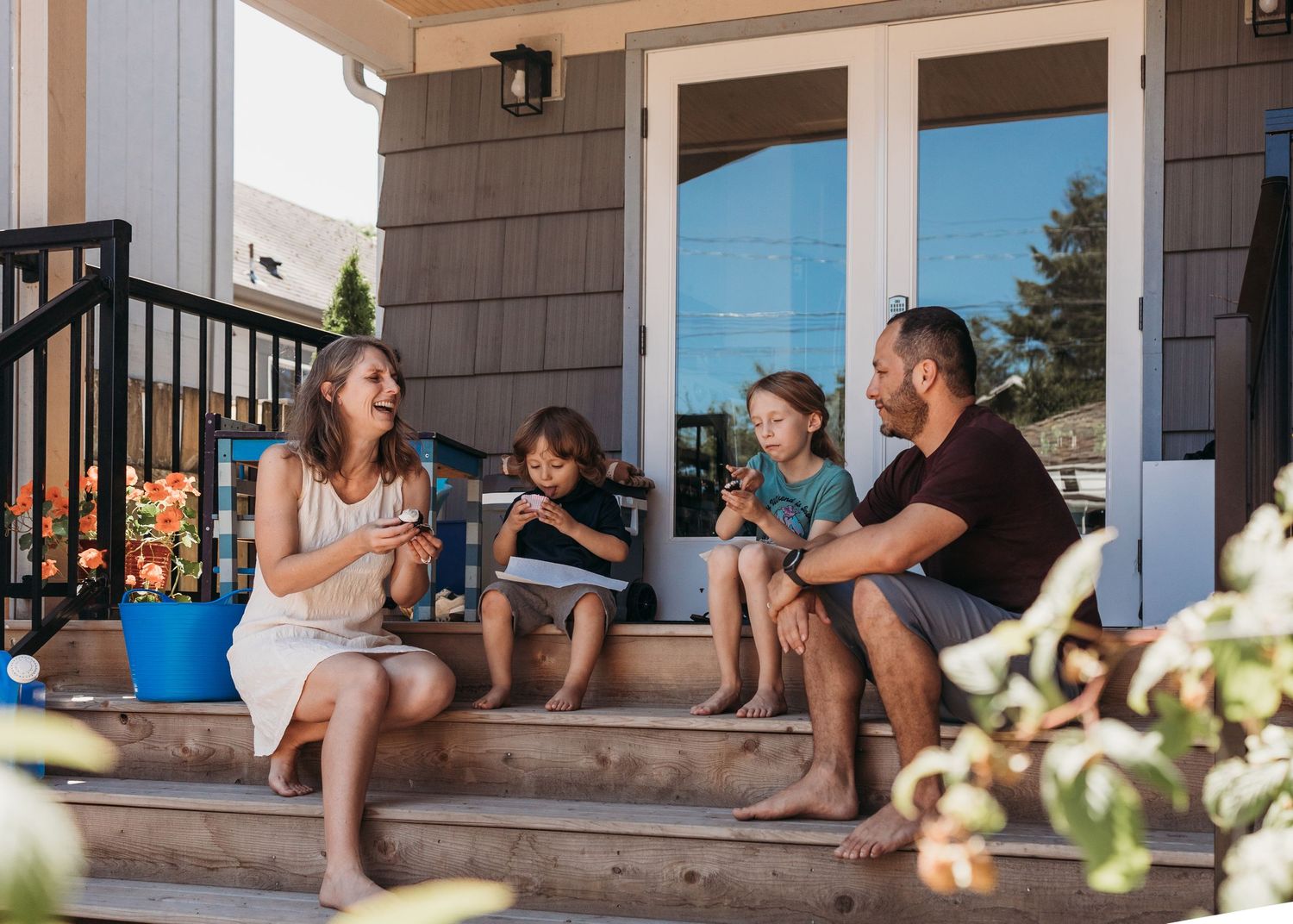 A family relaxing together on their front porch steps during a sunny afternoon, surrounded by potted flowers.
