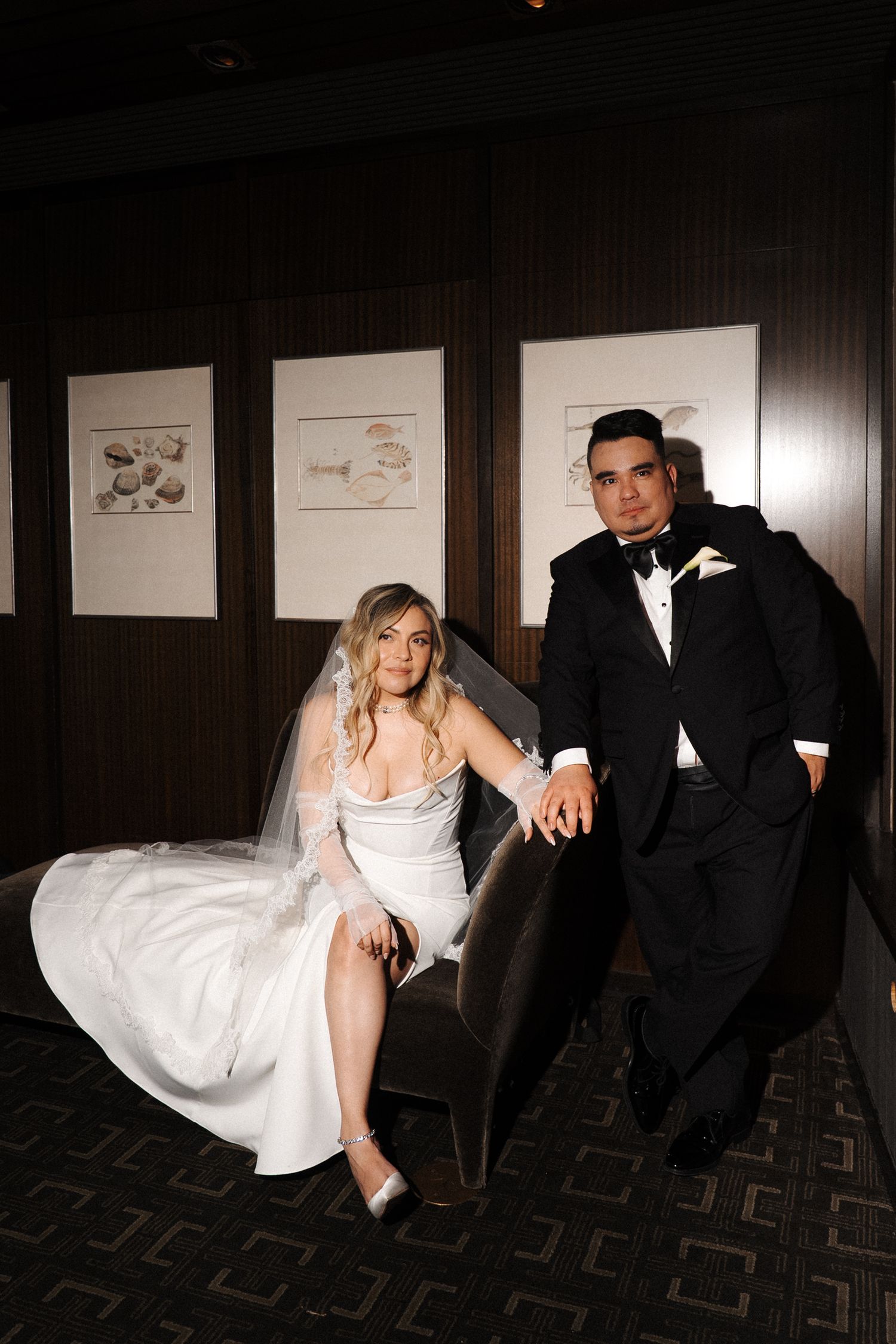 Wedding couple poses together in formal attire against dark wood-paneled wall with artwork.