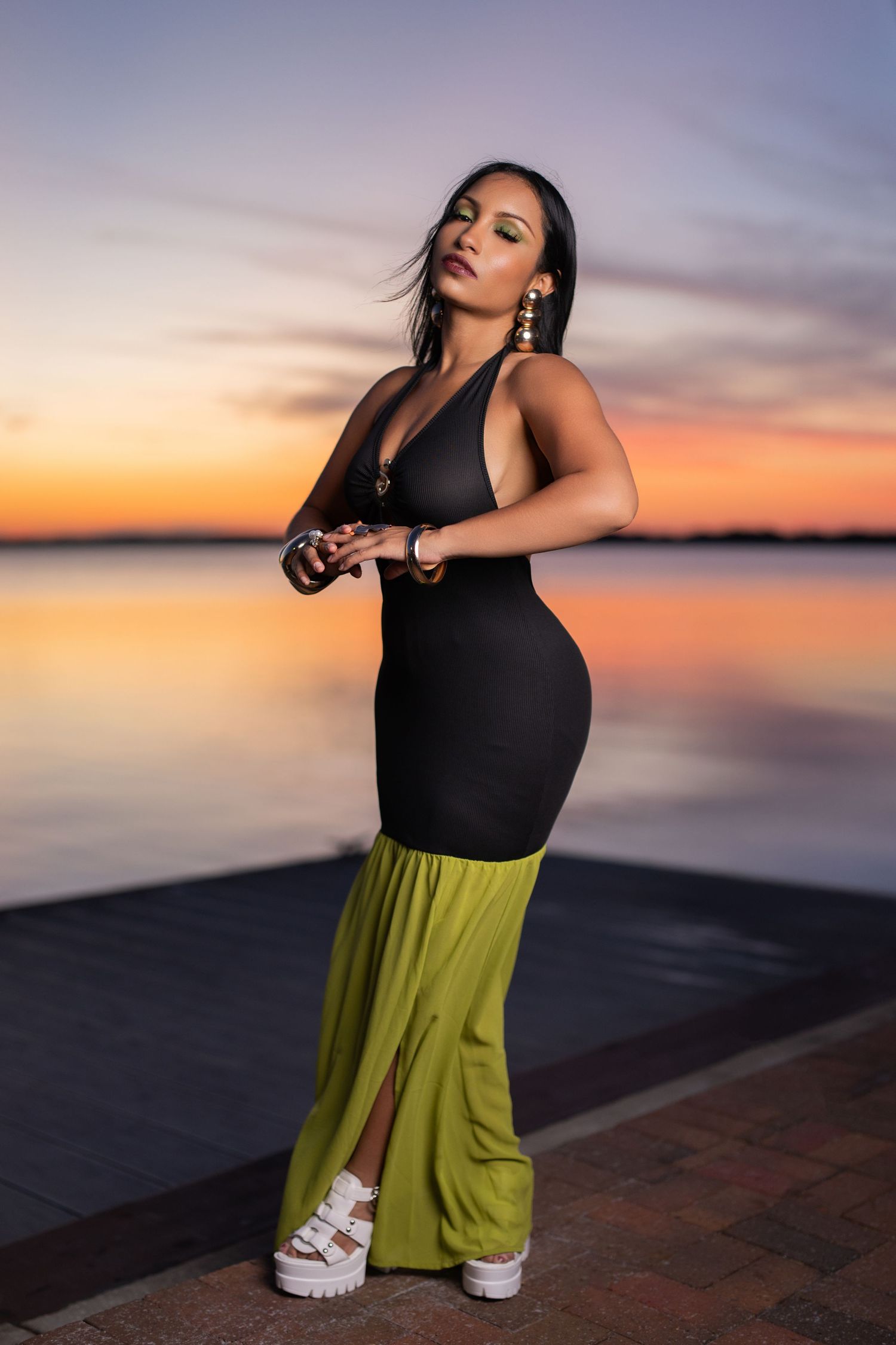Figure in black and lime green dress poses against vibrant sunset waterfront backdrop wearing white platform sandals.