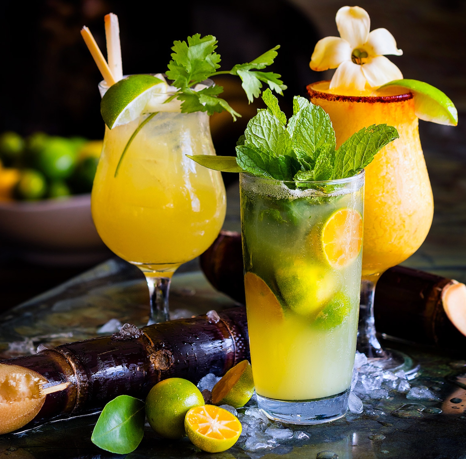 Three tropical cocktails garnished with citrus, mint and flowers served on dark background with fresh fruits.