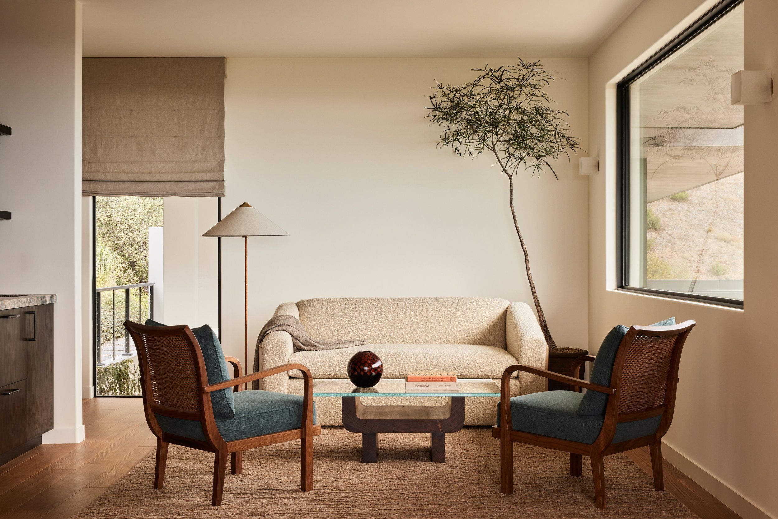 A modern minimalist living room with beige sofa, vintage leather chairs, coffee table and potted tree by large windows.