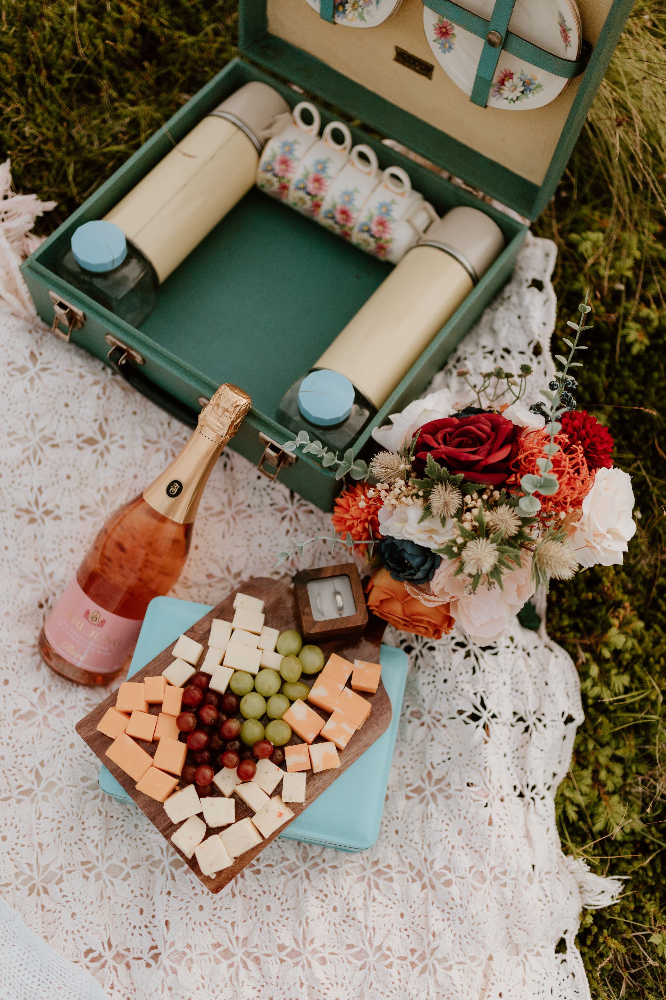 picnic elopement set up with an old fashioned picnic case, wedding bouquet, a bottle of champagne, and grapes and cheeses