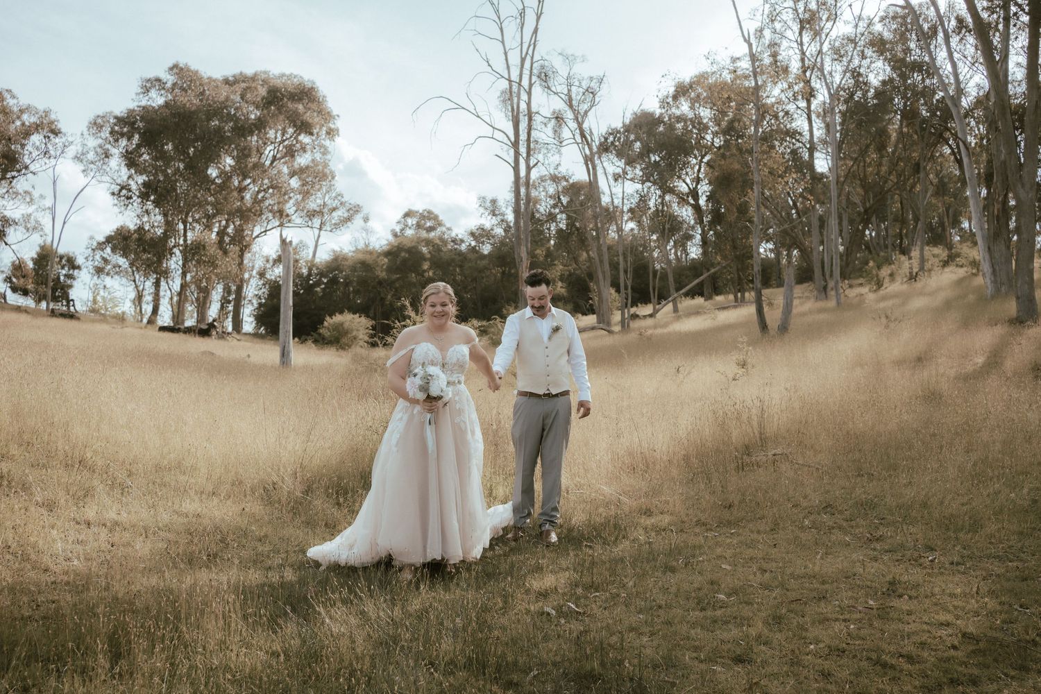 Couple in wedding attire embrace on a grassy hillside with rustic trees in the background at sunset.