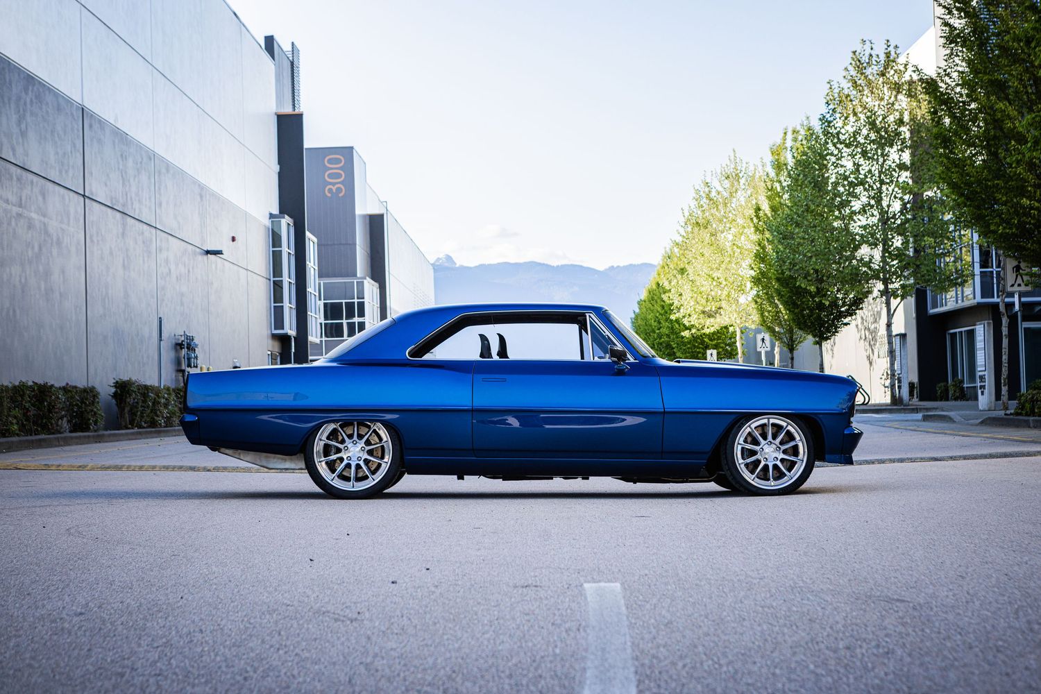 A sleek blue custom lowrider car with chrome wheels sits low to the ground in an urban setting.