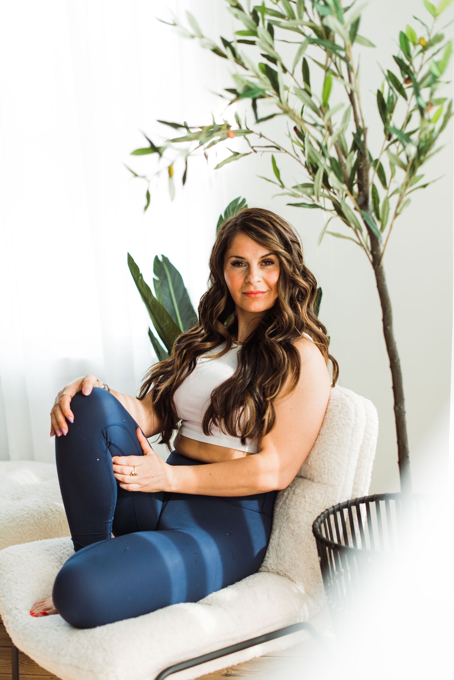 A woman in a white top lounges on a beige couch beside a decorative olive tree in a bright, minimalist setting.