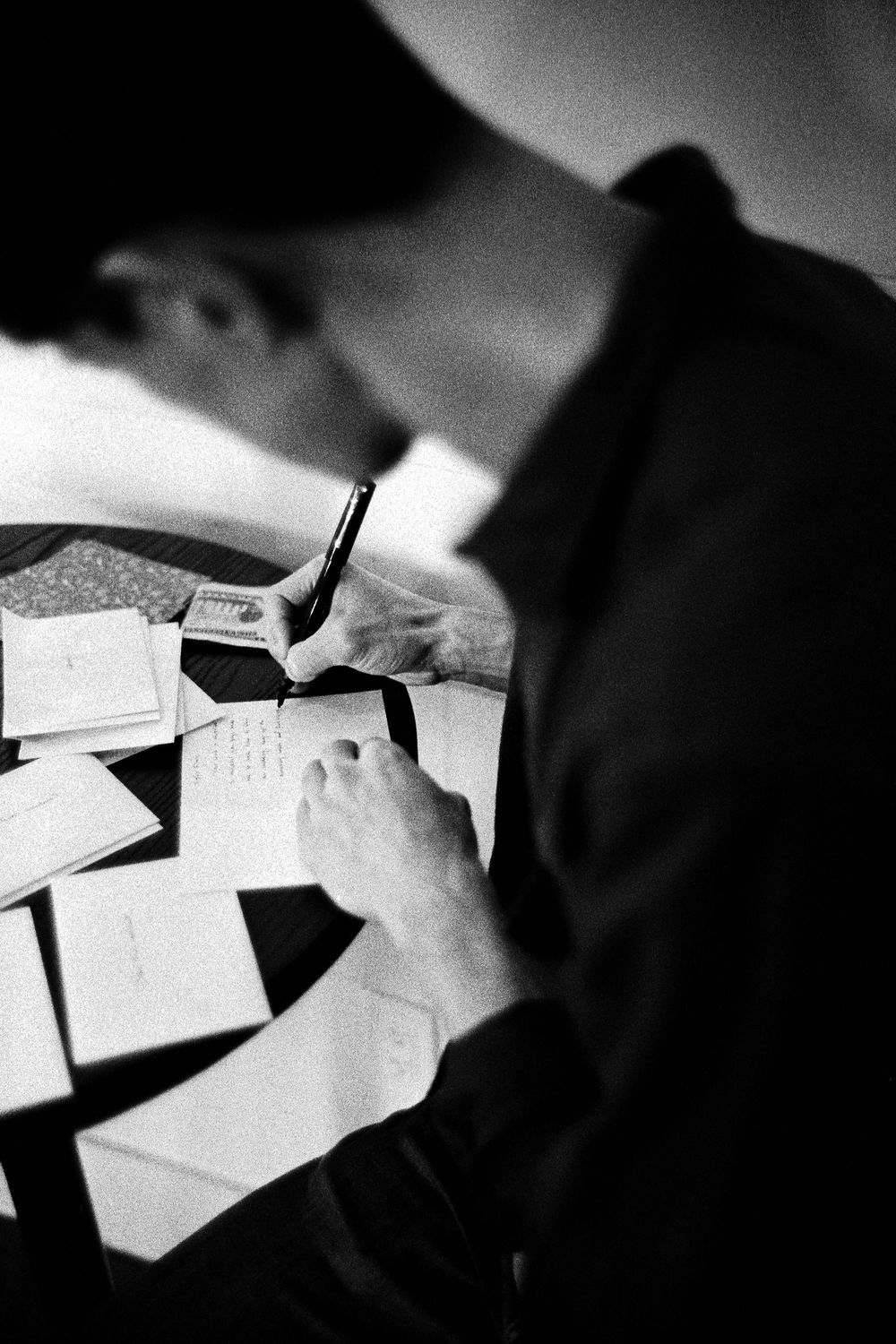 Black and white photo of a person in a dark cap writing notes on paper surrounded by documents on a table.