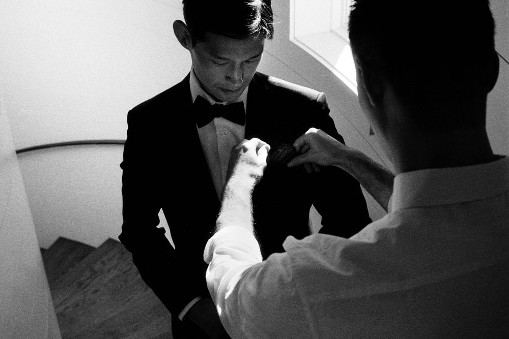 Black and white photo of groom in tuxedo being helped with boutonniere on staircase.