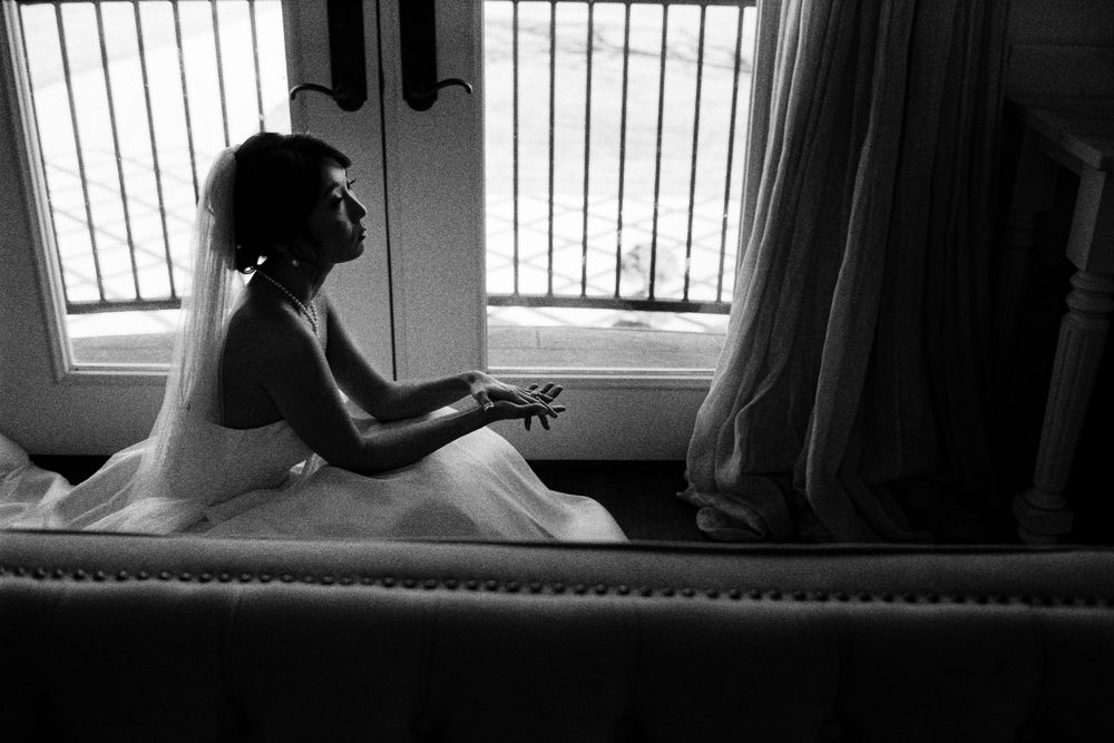 Black and white photo of bride in wedding dress sitting pensively by window light.