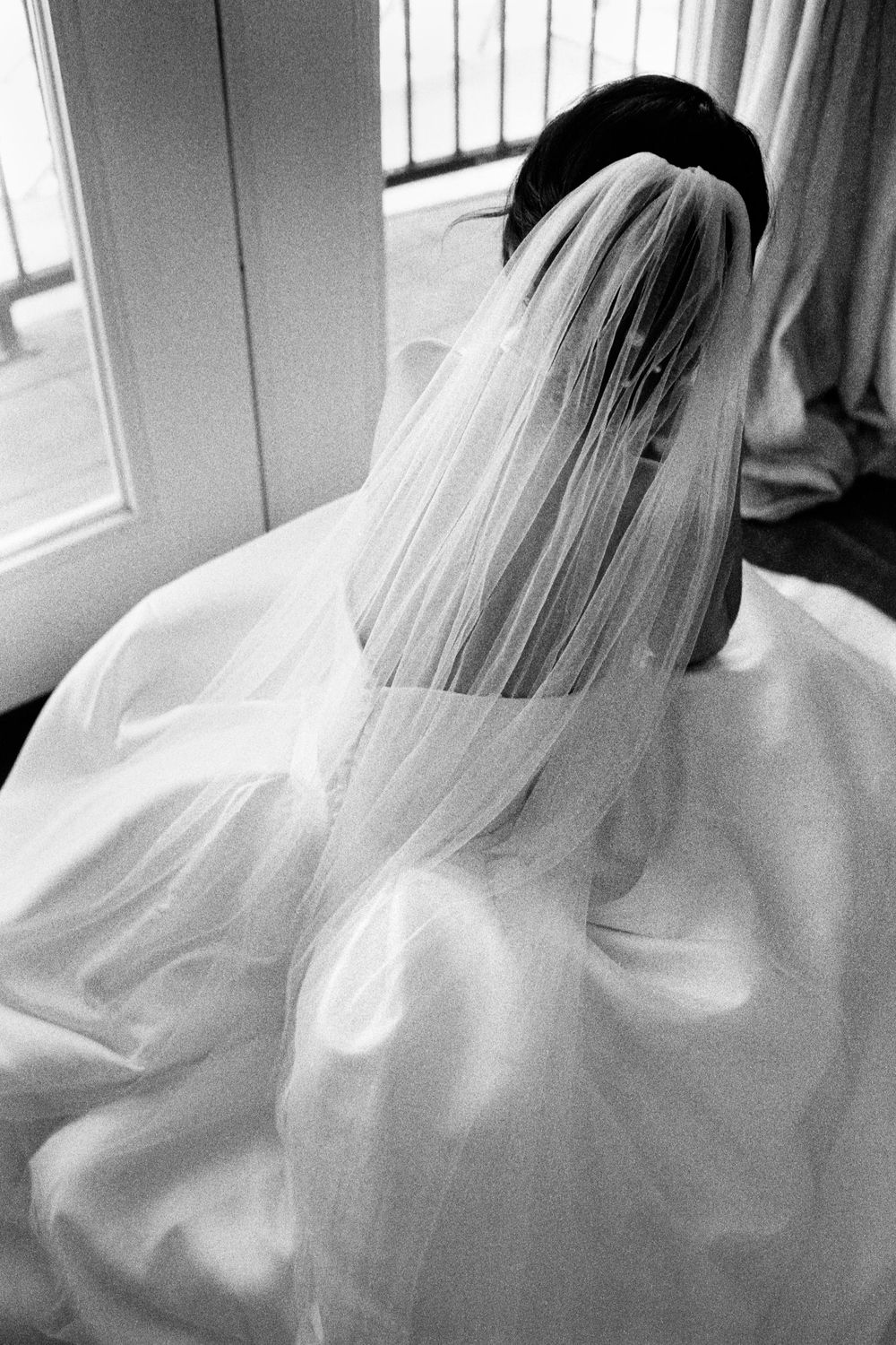 Black and white overhead photo of bride in white gown and veil seated by window.