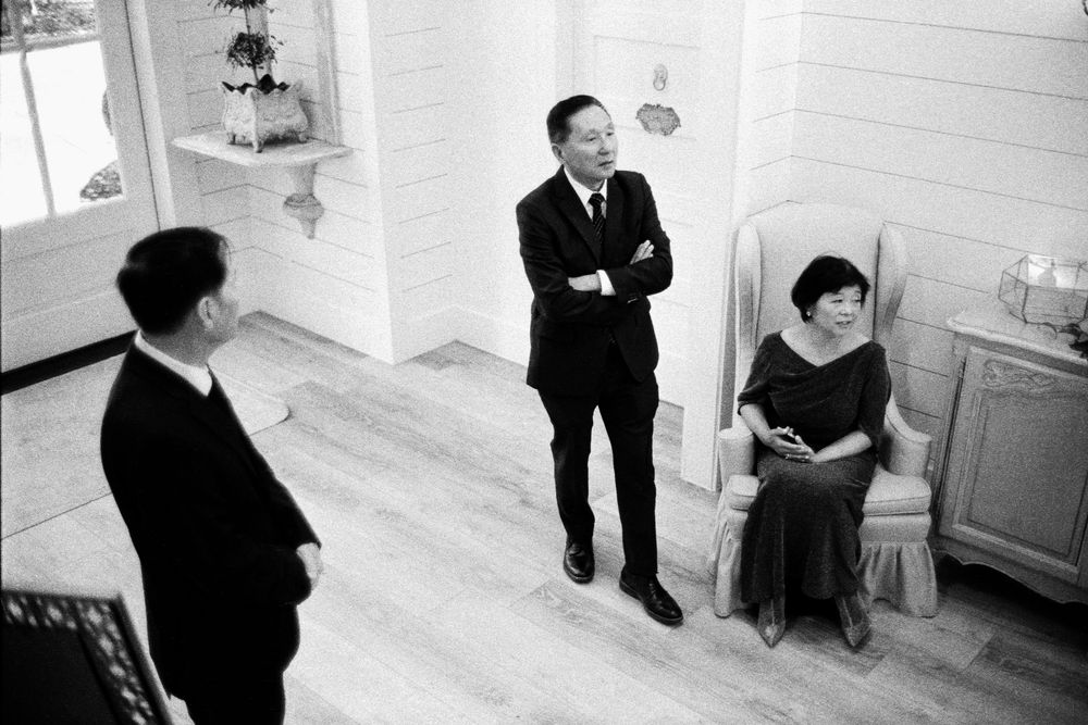 Black and white photo of three formally dressed guests conversing in a bright white-walled wedding venue room.