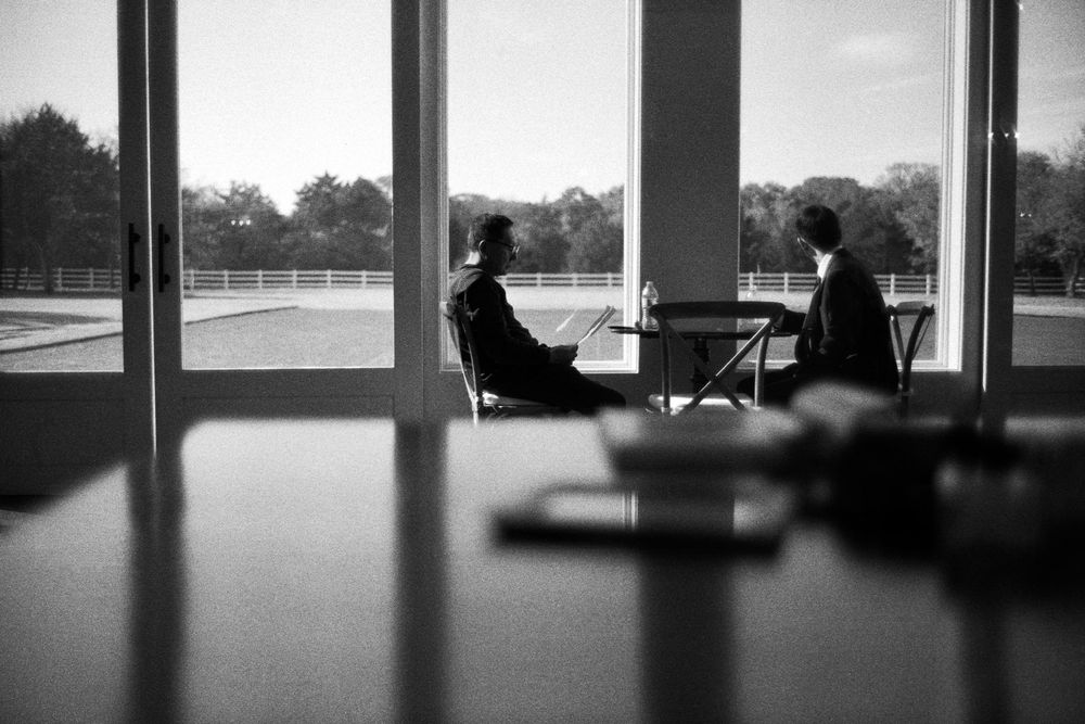 Black and white silhouette of two people seated at a table before large floor-to-ceiling windows outdoors.