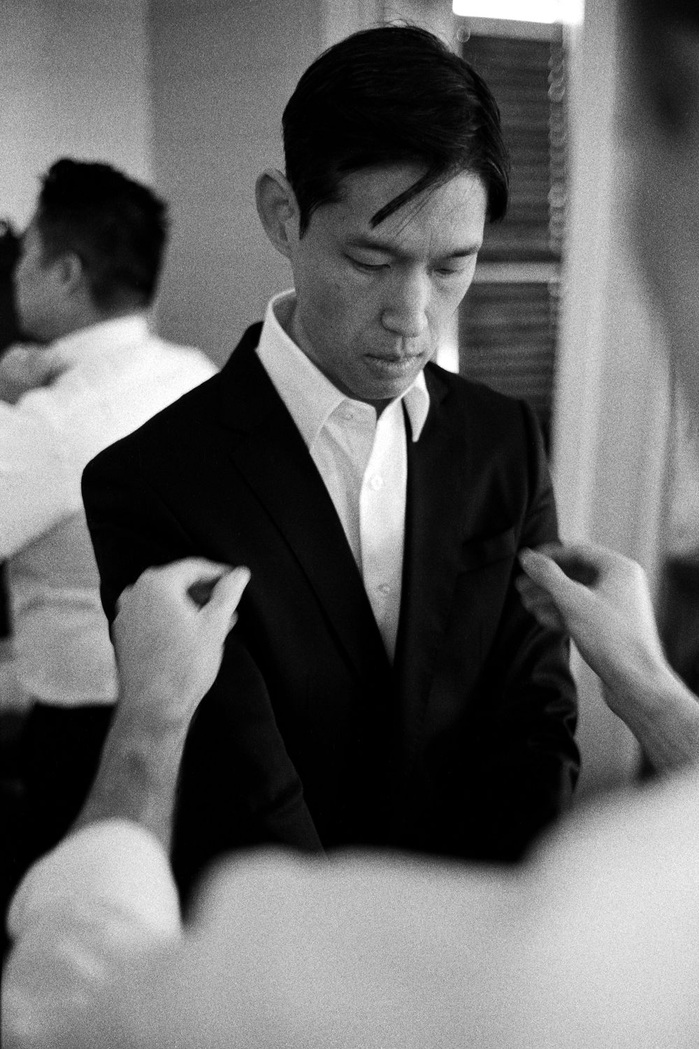 Groom being helped into black suit jacket by groomsmen during wedding preparations in black and white photo.