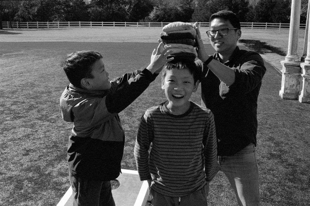 Black and white photo of two boys and a man playfully balancing a bag on a childs head outdoors.