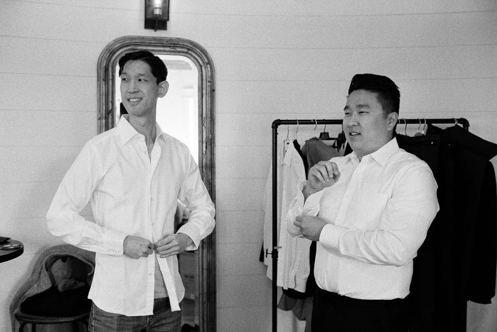 Two men in white dress shirts getting ready in a room with a mirror and hanging clothes.
