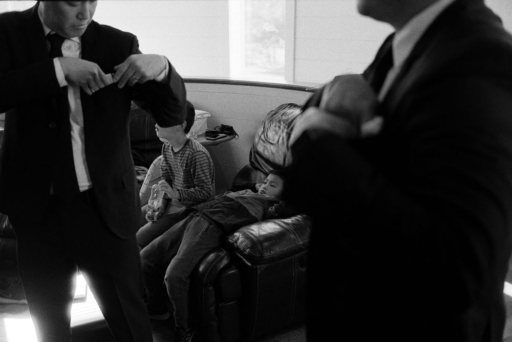 Black and white photo of groomsmen adjusting ties while a child sits on a couch nearby.