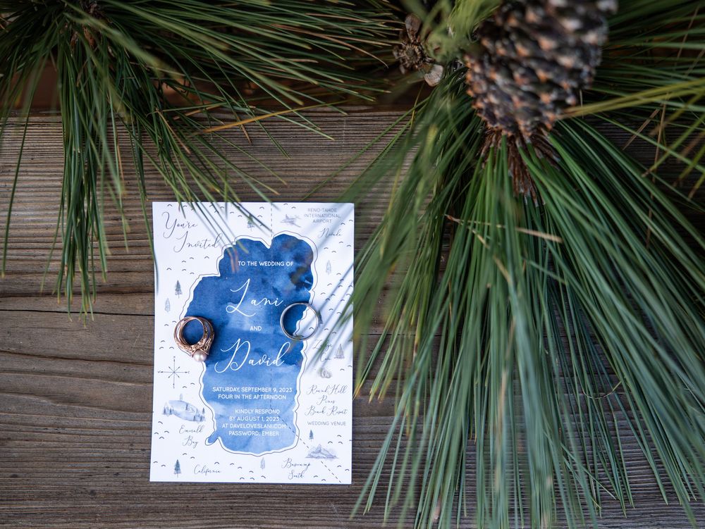 A blue wedding invitation card displayed among green pine needles and pine cones on a wooden surface.